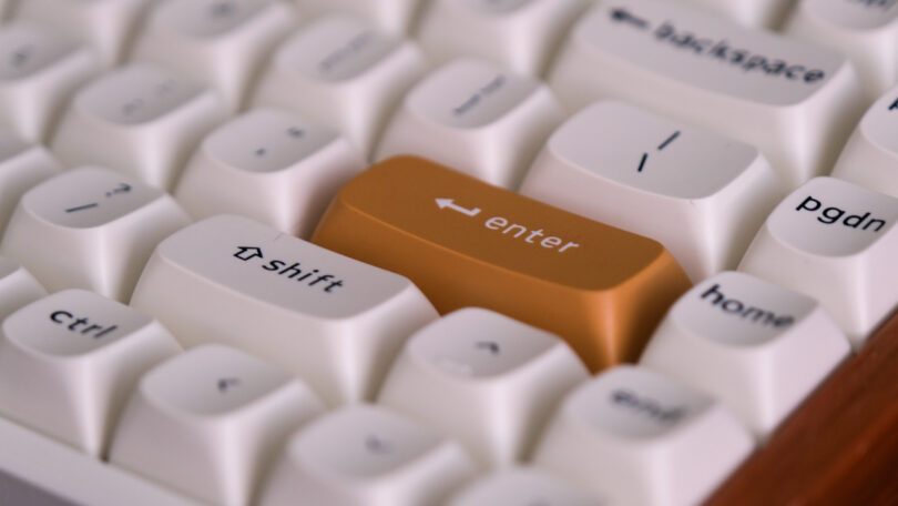 Close-up of a white keyboard with a prominently featured orange Enter key. Nearby keys include Shift, Ctrl, Backspace, and PgDn.