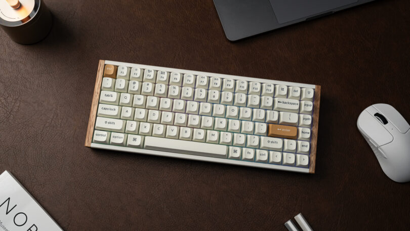 A compact mechanical keyboard with cream and gray keycaps sits on a brown desk surface next to a white computer mouse, a notebook, and a laptop.