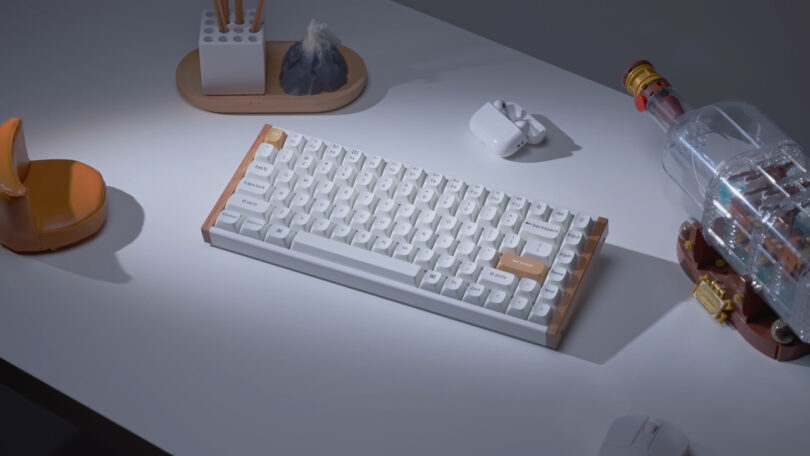 A white mechanical keyboard is on a desk, surrounded by a rubber duck, a wooden stand with a pen holder, an open case with earbuds, and a ship in a bottle.