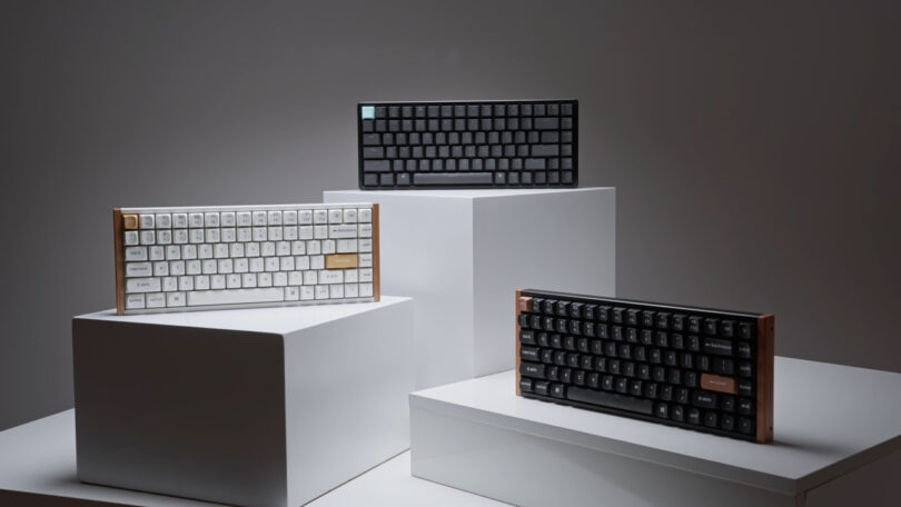 Three keyboards are displayed on white pedestals: one white and gold, one black, and another black with copper accents, all with mechanical keys. The backdrop is plain gray.