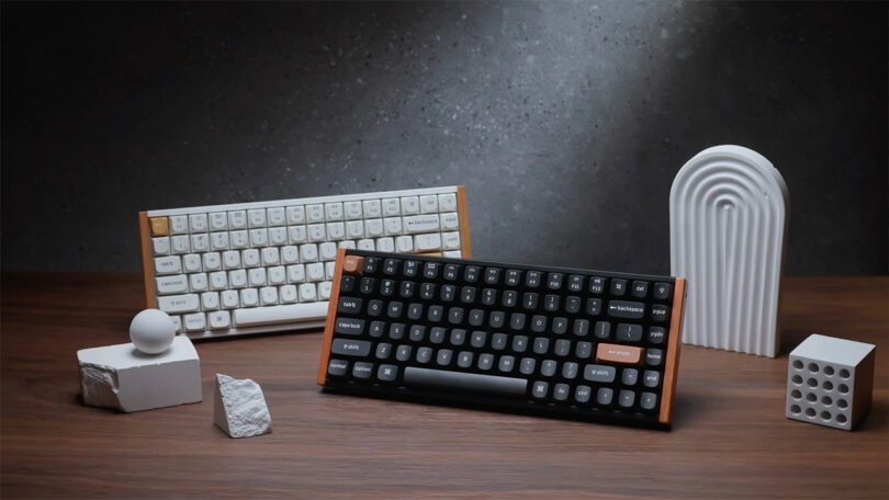 Two mechanical keyboards, including a sleek Keychron K2 HE, are displayed on a wooden surface—one with white keys and the other with black keys. Surrounding them are white geometric decor objects.