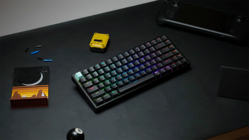 A Keychron K2 HE mechanical keyboard with RGB lighting sits on a dark desk next to various small objects, including a handheld gaming device, a yellow gadget, a small building block toy, and an 8-ball.