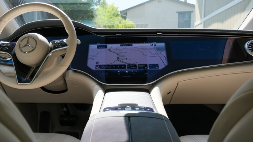 Interior front view of a modern car with a dashboard featuring a large touchscreen display, navigation map, and a Mercedes-Benz emblem on the steering wheel.