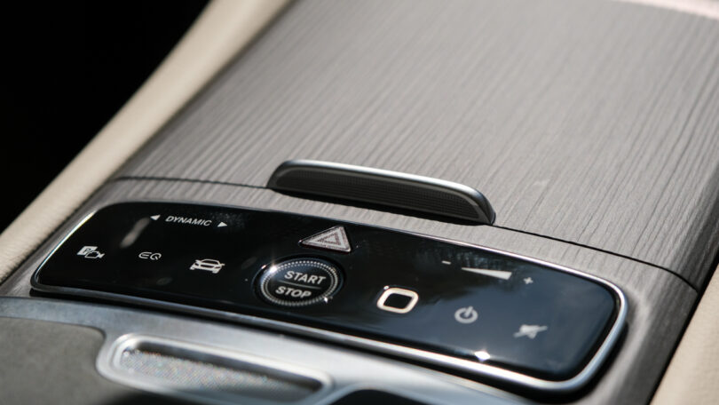 Close-up of a car's center console featuring a wood finish, various control buttons, including a "Start/Stop" button, and icon indicators for vehicle functions such as dynamic mode and hazard lights.