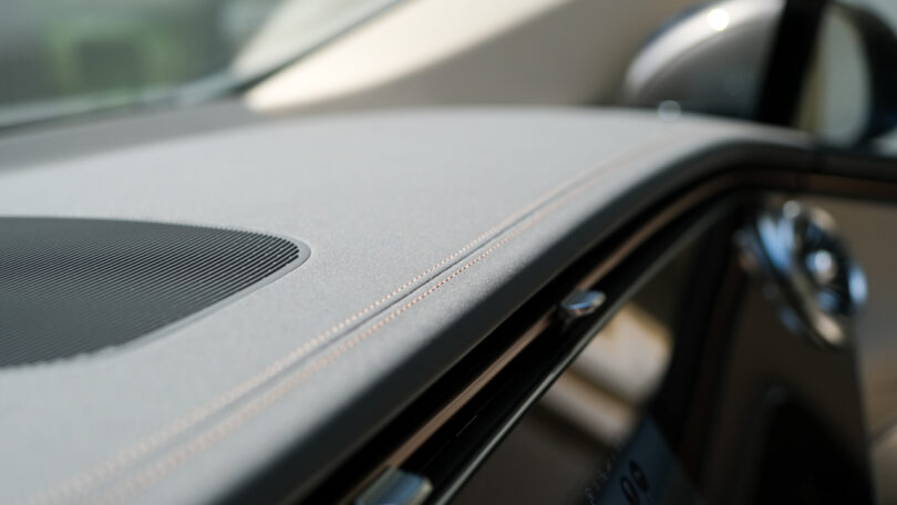 Close-up of a car dashboard featuring detailed stitching, part of the speaker grille, and a glimpse of the side mirror. The image highlights the fine craftsmanship and interior design details of the vehicle.