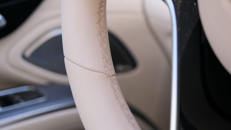 Close-up of a beige leather car steering wheel showing detailed stitching, with parts of the interior and control panel blurred in the background.