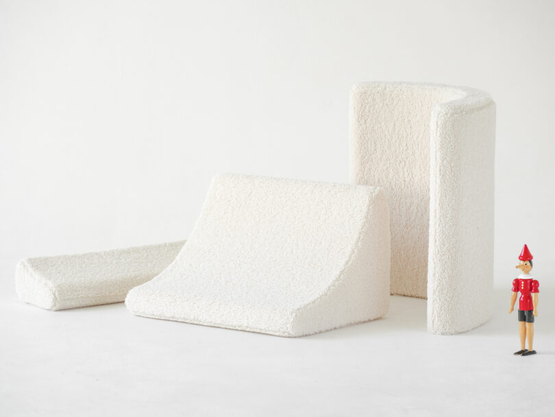 Three different shapes of white textured cushions are arranged on the floor with a small wooden toy figure in a red outfit standing next to them