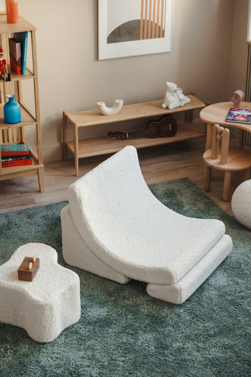 A cozy reading nook includes a low padded chair, ottoman, wooden side table with magazines, bookshelf with colorful items, framed wall art, and a small guitar on the floor