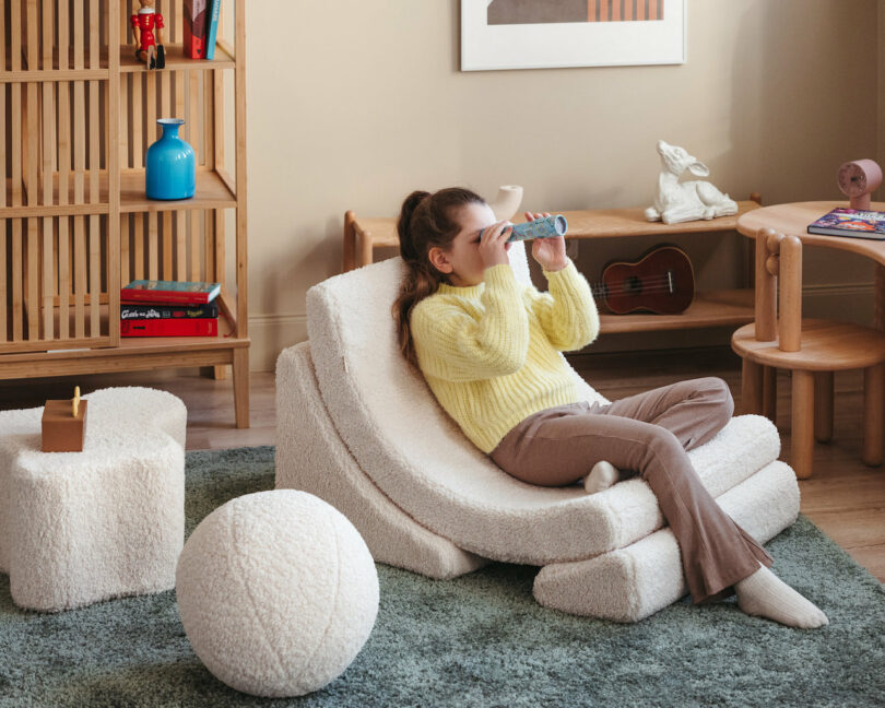 A child sits on a white cushioned chair in a cozy room, looking through a handheld paper tube. The room has a wooden shelf with items, small tables, and a large ball on a green carpet