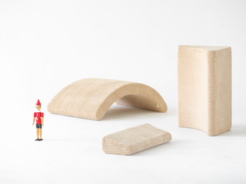 Wooden Pinocchio doll standing next to three curved and rectangular beige fabric-covered objects on a white background