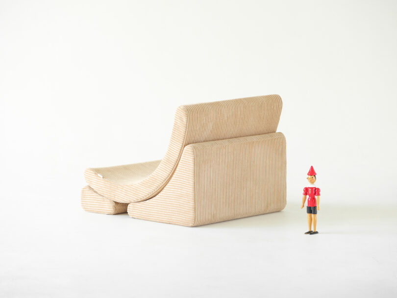 A tan, corduroy sectional sofa positioned on a white background. A small wooden toy figure stands to the right of the sofa