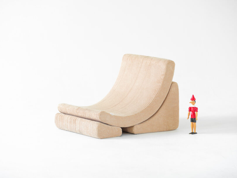 A beige, curved, modern chair with a small wooden figure wearing a red outfit and hat standing beside it. The background is plain white