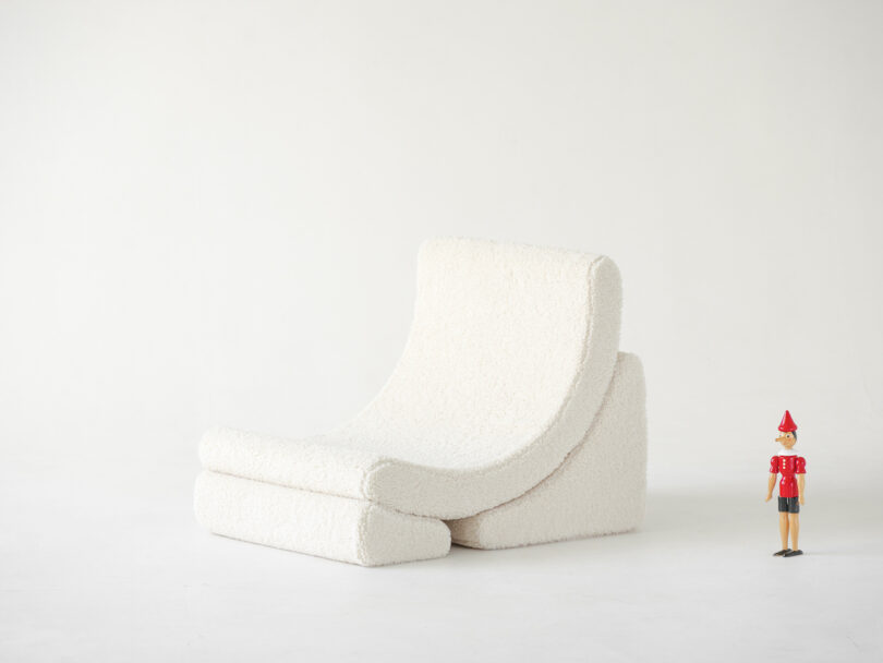 A plush, white, modern lounge chair with a textured fabric is set against a plain background. A small wooden figurine in a red outfit and hat stands next to the chair