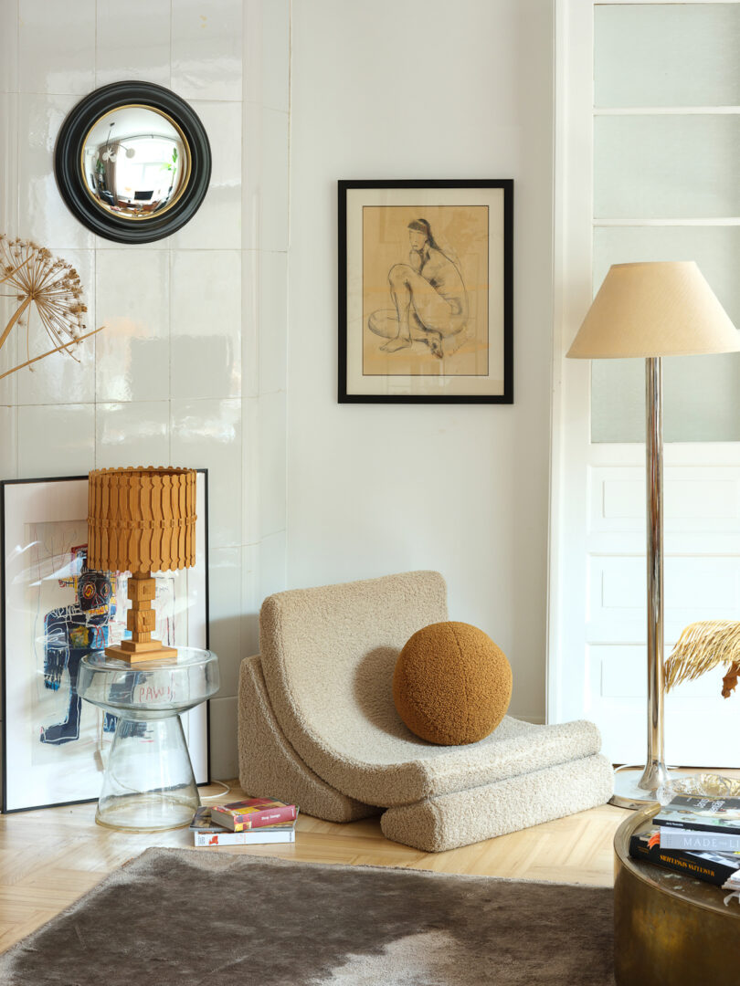 A cozy interior corner features a beige lounge chair with an orange cushion, a transparent side table holding a woven lamp, framed artwork on the wall, and a carpet on the floor