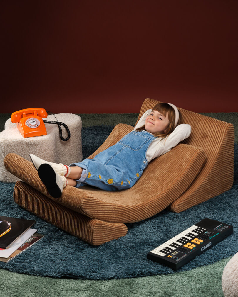 A child wearing a denim overall rests on a corduroy lounge chair with legs crossed. Beside are a rotary phone, a keyboard, and books on a carpeted floor