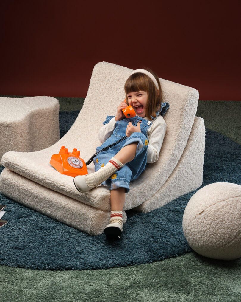 A child in denim overalls and white sweater sits on a plush chair, holding an orange rotary phone to their ear, with a matching orange receiver resting on their lap