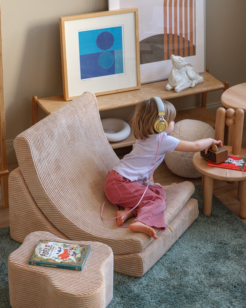 A child wearing headphones leans on a textured chair, reaching towards a wooden toy on a small table. Children's books are on the floor, and artwork is displayed in the background