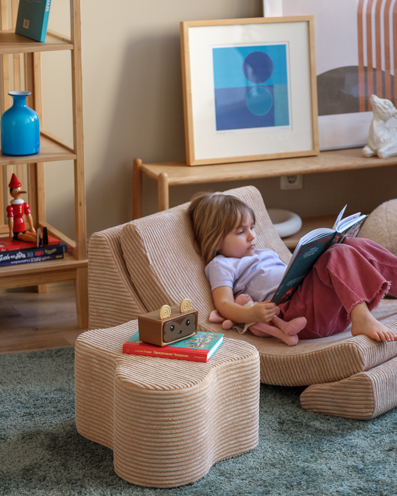 A child sits in a beige curved armchair, reading a book. Nearby is a matching ottoman with a toy and two more books on it. Shelves and framed artwork are visible in the background