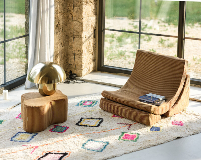 A modern room with a low wooden chair, a matching table, and a metallic lamp. A colorful patterned rug covers the floor, and large windows provide natural light. Books are placed on the chair