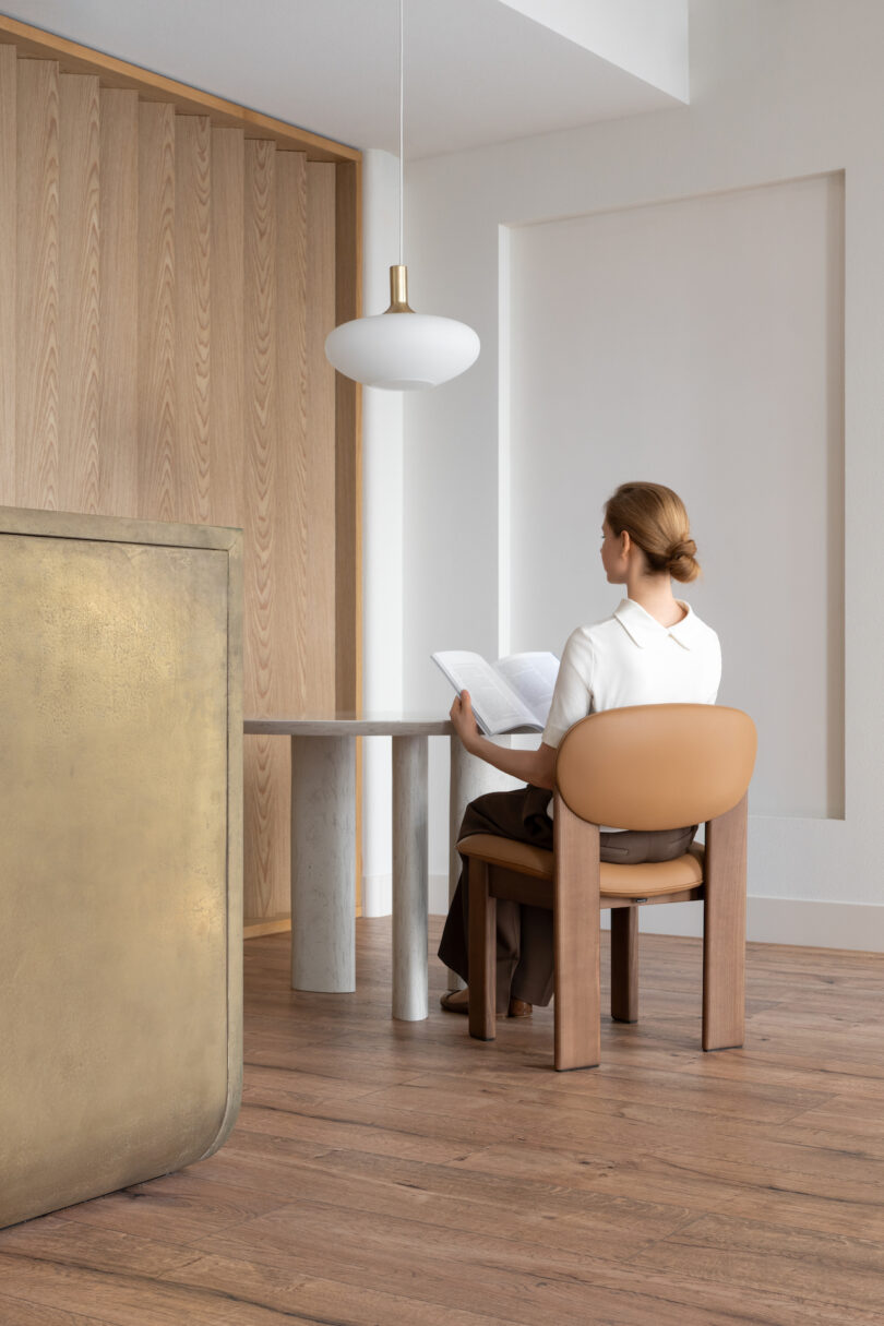 A person with tied-back hair sits on a light brown chair, reading a magazine at a round table in a minimalist room with wooden flooring and walls. A white pendant lamp hangs above the table