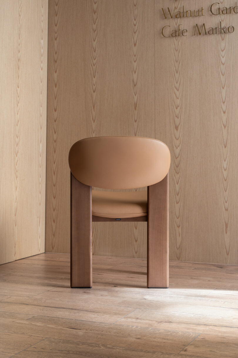 A brown chair with a curved backrest and solid wooden legs is positioned against a light wood-paneled wall with text partially visible in the upper right corner