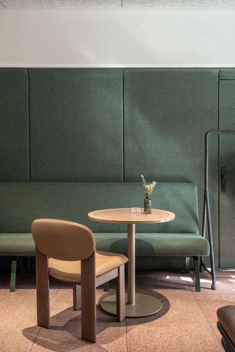 A small round table with a vase of dried flowers, one tan chair, and a green padded bench against a green wall in a minimalist room