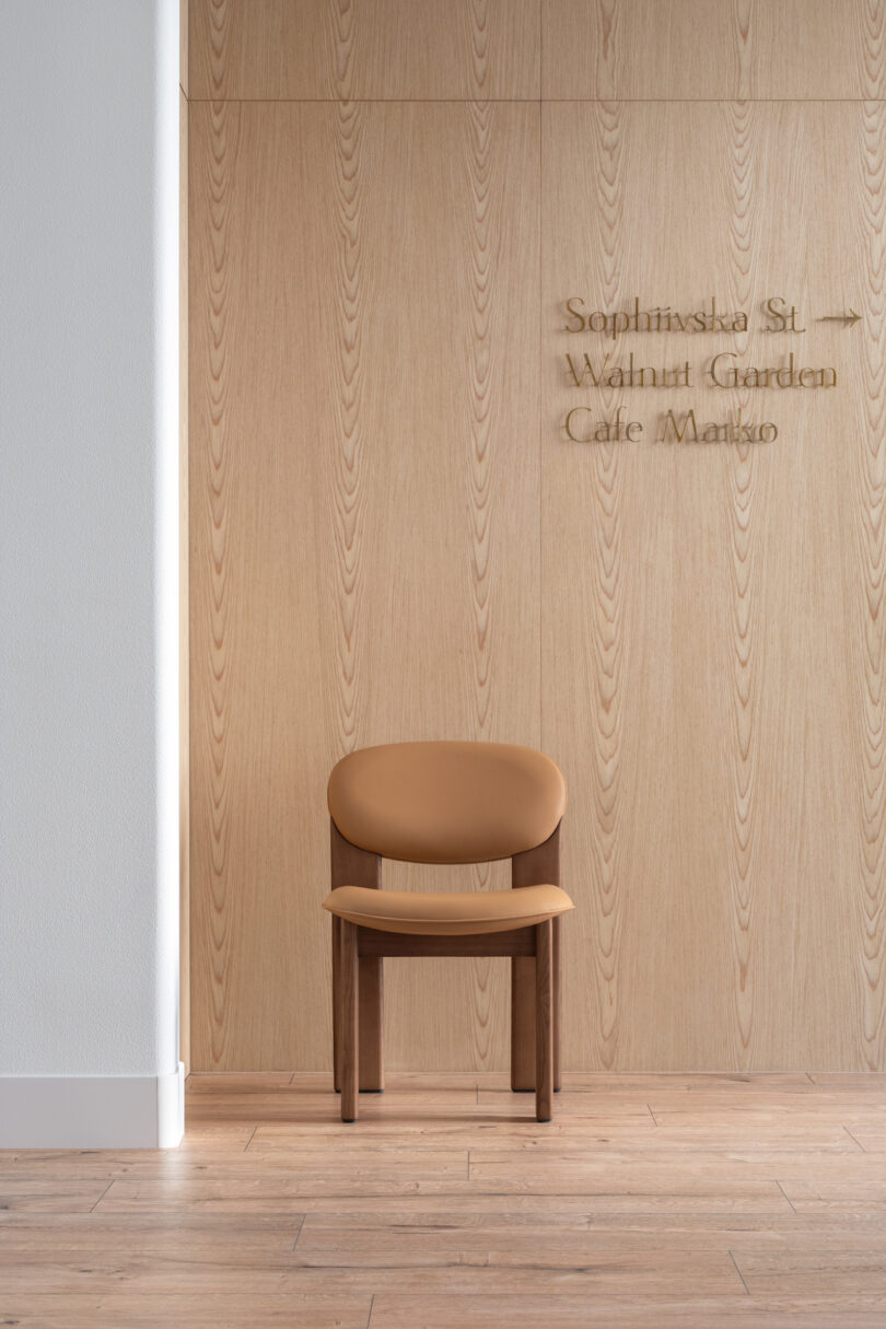 Minimalist wooden chair set against a light wooden panel wall, with carved text on the wall listing "Sophinska St. Walnut Garden Cafe Margo." 