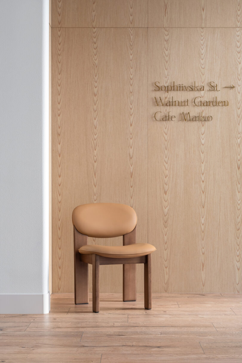 A tan chair is placed against a light wood wall with text listing "Sophinska St., Walnut Garden, Cafe Margo." The floor is made of light-colored wood, and a white wall is visible on the left