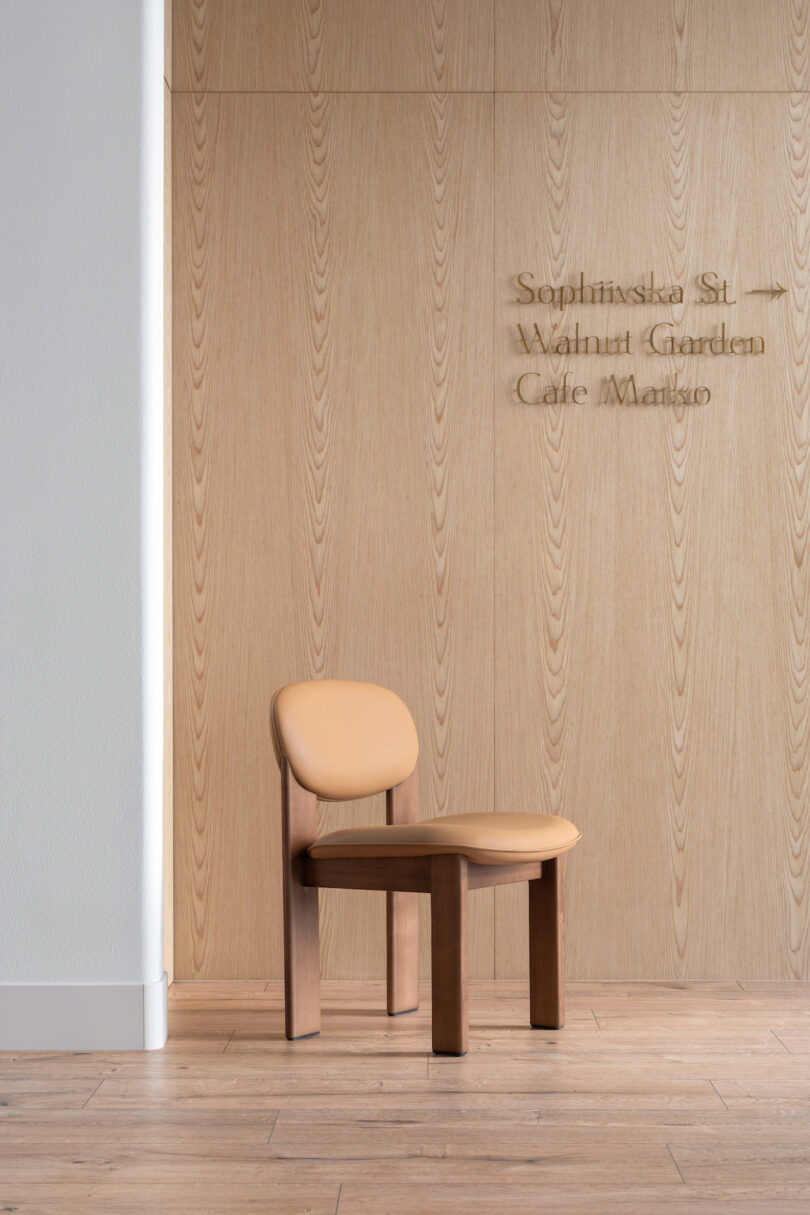 A tan wooden chair with a padded seat and backrest is placed against a light wood panel wall that has text on it, near a white corner wall