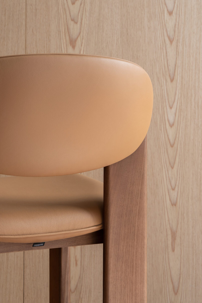 Modern chair with a curved backrest and cushioned seat, upholstered in beige leather. The chair has wooden legs and is placed against a wooden panel wall with a similar wood grain pattern