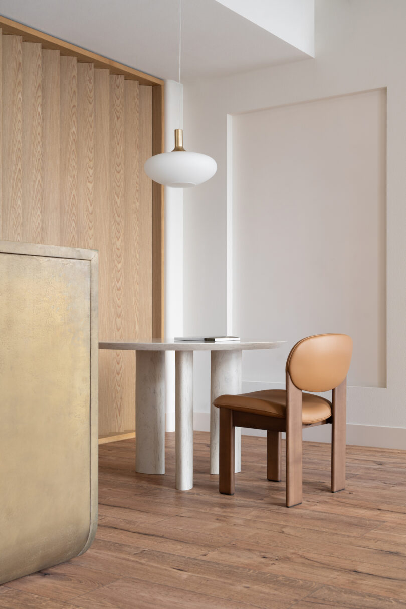 A modern dining area featuring a small round table, a single brown chair, a hanging white pendant light, and a wooden accent wall. A partially visible beige structure is in the foreground