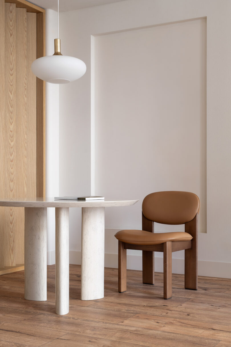 Minimalist interior with a round marble table, a leather chair, a hanging pendant light, and wooden flooring