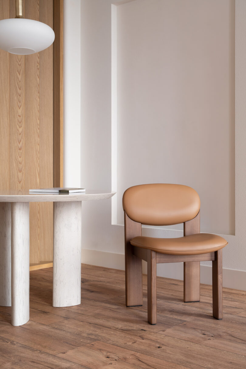 A wooden chair with a tan cushion is placed beside a white, round table with an open book. The setting features wooden flooring and a minimalist light fixture