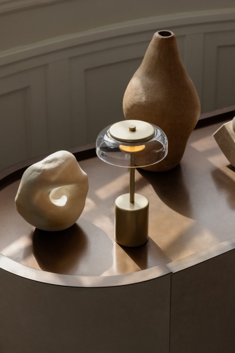 A modern table lamp with a clear glass shade stands on a brown table along with two abstract ceramic sculptures and a large, rustic vase