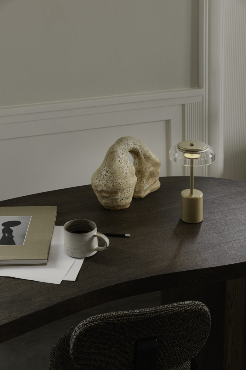 A dark wood desk with an abstract stone sculpture, small lamp, coffee cup, pencil, and books, set against a neutral wall, with part of a cushioned chair visible in the foreground