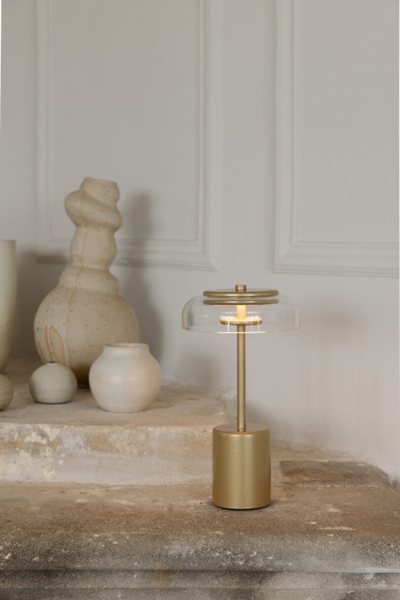 A gold table lamp with a glass shade is placed on a stone shelf next to various ceramic vases in neutral tones