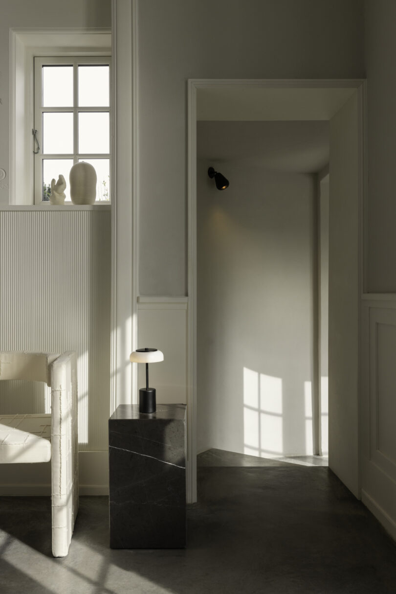 Minimalist interior featuring a white armchair, small black side table with a lamp, and a window casting light on the walls and floor