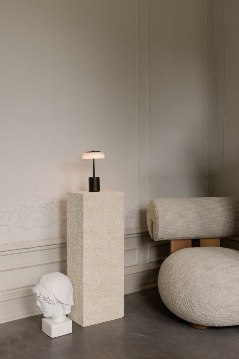 A minimalist room with a cushioned chair, a stone pedestal holding a modern lamp, and a small sculpture of a classical head. The wall features subtle decorative molding