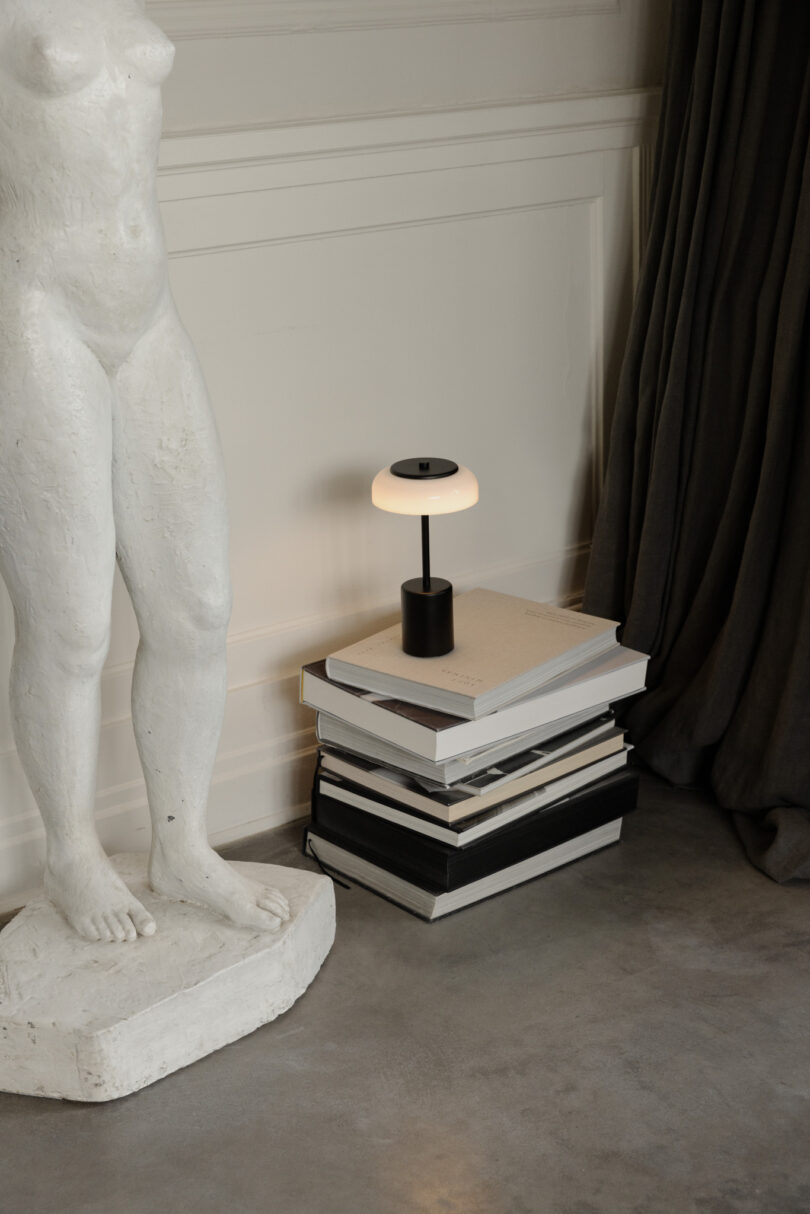 A stack of books serves as a table for a small modern lamp. A white sculpture of a standing human figure is placed to the left, and a dark curtain hangs in the background