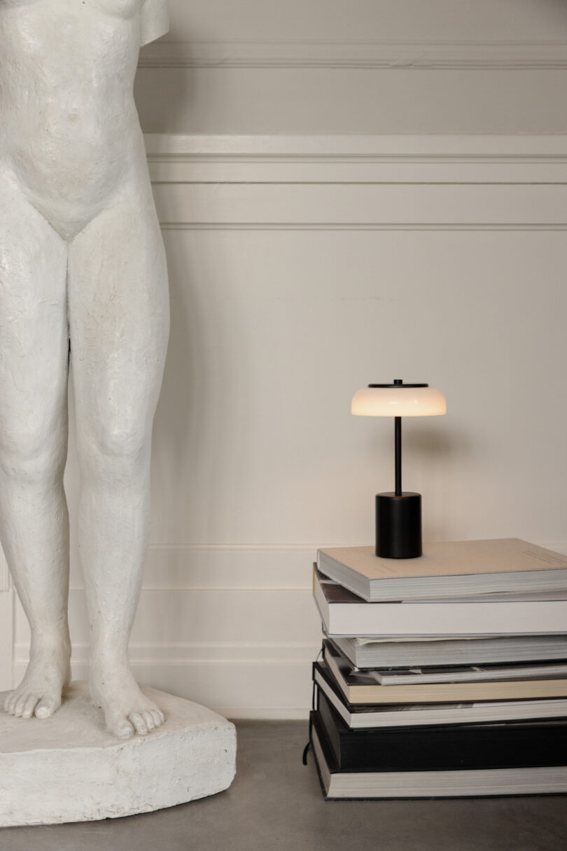 A white statue stands against a wall next to a stack of books topped with a small black lamp