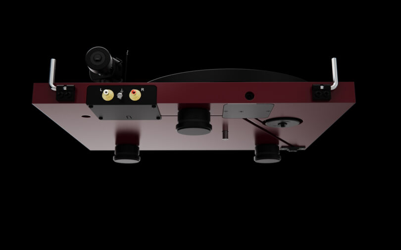 Bottom view of a turntable in a red housing, showing its various components including gold-plated RCA jacks, a belt drive mechanism, and adjustable feet against a black background.