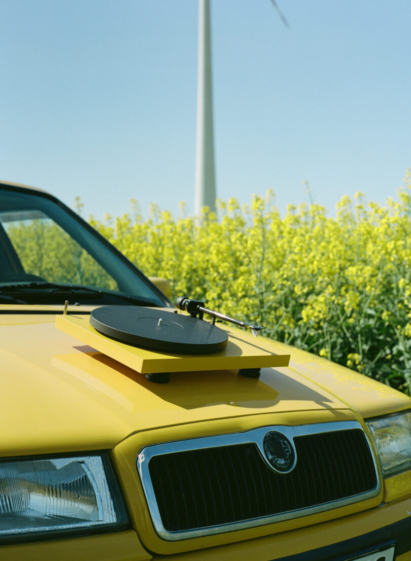 Yellow car with a turntable on its hood, parked in front of a field of yellow flowers and a wind turbine in the background.