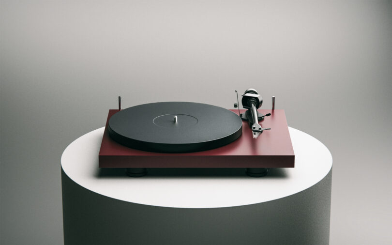 A red turntable with a black platter and tonearm is displayed on a cylindrical white pedestal against a gray background.