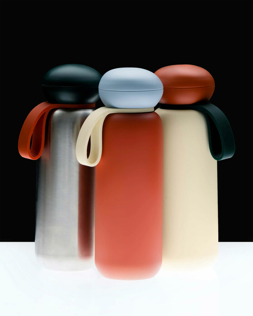 Three reusable bottles with round caps and strap handles are lined up. The Sunnies Flask is metallic with a black cap, the next is red with a light blue cap, and the third is beige with an orange cap.