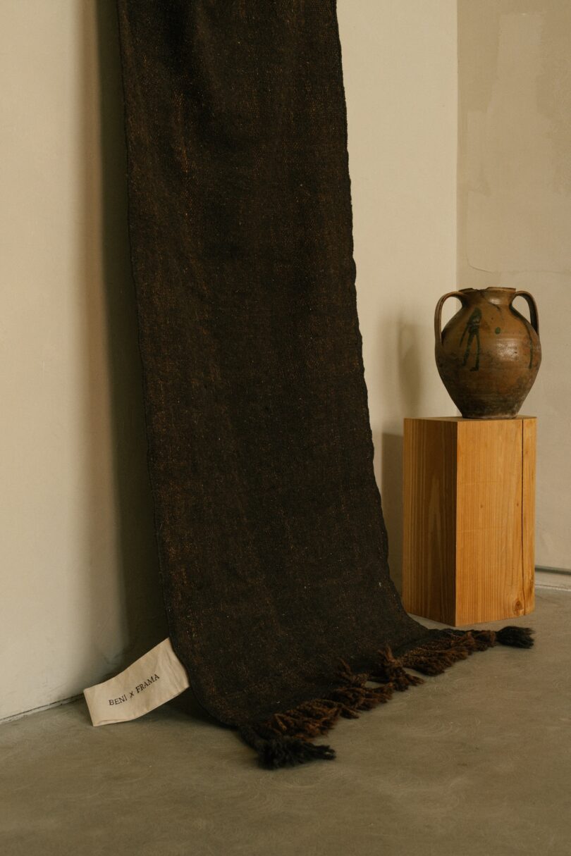 A dark Beni rug from the Terrain collection draped against a wall, paired with a rustic pottery piece on a wooden pedestal