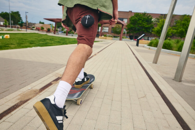 Person wearing brown shorts and white socks skateboarding in a paved outdoor area, with a round patch on their shorts. Buildings and green area visible in the background.