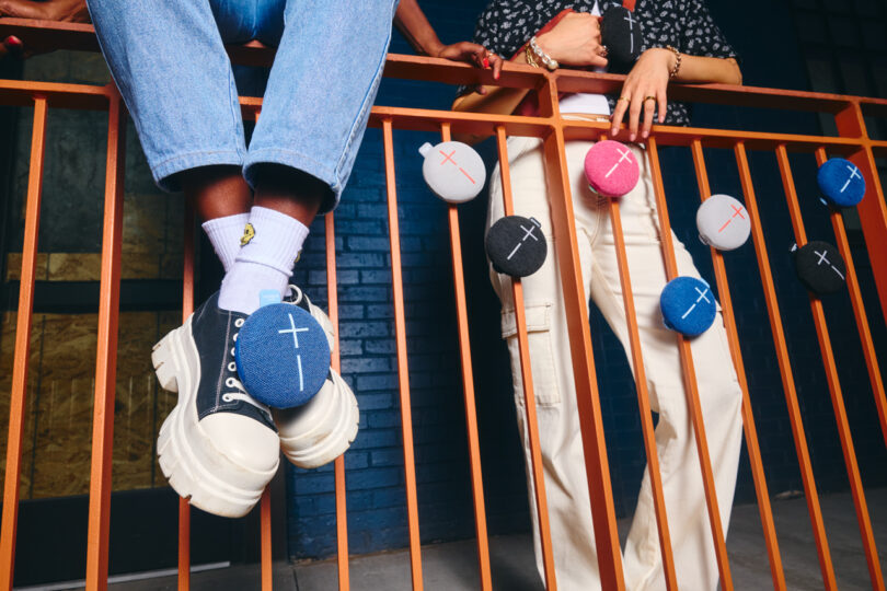 Two people standing behind an orange metal railing with multiple colored Bluetooth speakers clipped to it. Visible parts include legs, footwear, and accessories.