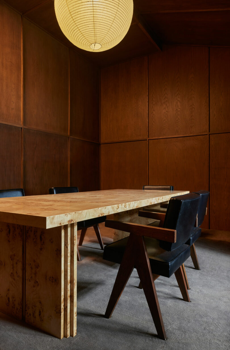 A minimalist conference room features a large, light wood table with three black chairs on each side, set against dark wood paneled walls, and lit by a single round paper pendant light.