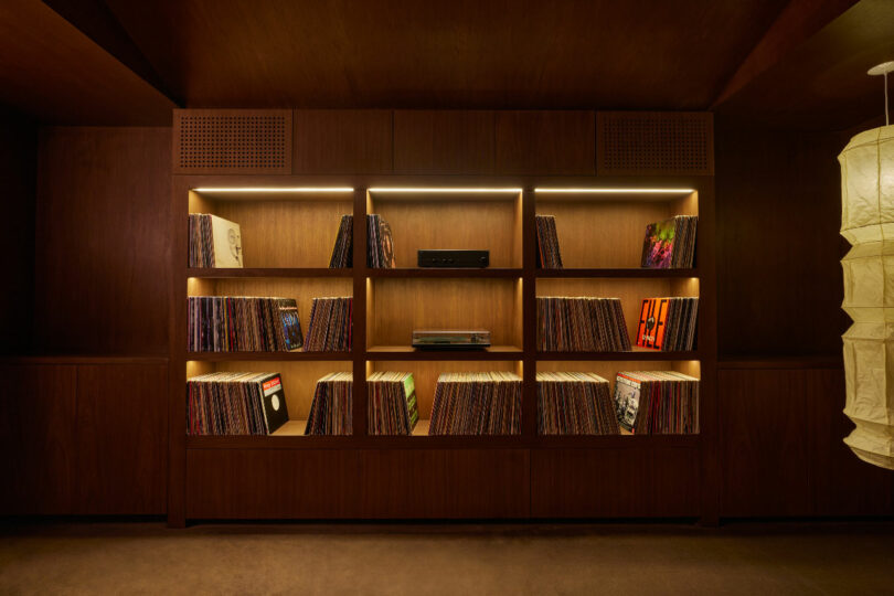 A wooden bookshelf with integrated lighting, filled with vinyl records and electronic equipment, stands in a dimly lit room. A white paper lantern is visible on the right side.