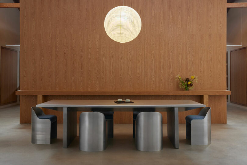 Modern dining room with a long metallic table, matching metallic chairs, a wooden wall backdrop, a spherical pendant light, and a flower arrangement on a side shelf.